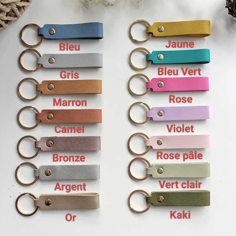 May include: A collection of fourteen rectangular keychains in various colors, each featuring a gold-tone metal ring and a central gold-tone rivet. The colors include blue, gray, brown, camel, bronze, silver, gold, yellow, teal, pink, violet, pale pink, light green, and olive.