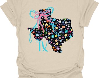 May include: A beige t-shirt with a floral Texas outline, a bow, and the letters "TX" in turquoise. The Texas outline is filled with colorful flowers. The bow is pink and turquoise.