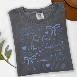 May include: A gray t-shirt with the text "Just One More Chapter" in blue script, surrounded by blue illustrations of books, hearts, bows, and a teacup. The shirt has a crew neck and a tag that says "Comfort Colors".