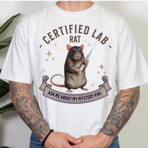 May include: A white t-shirt featuring a graphic of a brown rat holding a syringe. The text reads "CERTIFIED LAB RAT" in an arc above the rat and "ASK ME ABOUT MY MYSTERY VIAL" on a banner below. Small star icons surround the rat on the shirt.