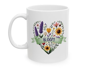 Może przedstawiać: Biały kubek ceramiczny z kwiatowym wzorem w kształcie serca i napisem "BLOOM" na zielonej wstążce. Serce jest wypełnione różnymi kwiatami w odcieniach fioletu, żółtego, czerwonego i pomarańczowego.