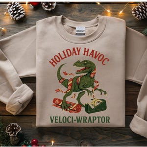 Puede incluir: Sudadera beige con un gráfico de dinosaurio verde envuelto en cinta navideña, sosteniendo cinta adhesiva y un regalo envuelto. El texto dice "HOLIDAY HAVOC" arriba y "VELOCI-WRAPTOR" abajo.