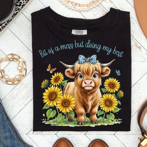 May include: Black t-shirt featuring a cartoon Highland cow with a blue bow, surrounded by sunflowers and butterflies. The text "Bit of a mess but doing my best" is written in blue script across the top. A cute and colorful design.