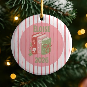 Può includere: Un ornamento rotondo in ceramica con sfondo bianco e strisce verticali rosse. Un cerchio rosa centrale mostra tre libri illustrati in rosso, rosa e verde, con il nome ELOISE e l'anno 2026 stampati in testo di colore verde.