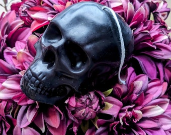 May include: A black skull candle sits on a bed of purple and pink flowers. The candle has a white wick and is made of wax.