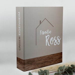 Könnte beinhalten: Ein brauner Ordner mit einem Holzboden. Der Vorderdeckel hat einen hellbraunen Hausumriss mit dem Text "Familie Ross" und die Worte "Unser Eigenheim" auf dem Rücken.