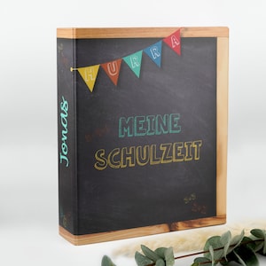 Könnte beinhalten: Ein schwarzer Ordner mit einem Holzrahmen und einem Kreidetafel-Design. Die Kreidetafel hat bunte Girlanden mit dem Wort "HURRA" darauf geschrieben. Der Text "MEINE SCHULZEIT" ist in bunter Kreide auf die Kreidetafel geschrieben. Der Name "Jonas" steht auf der Seite des Ordners.