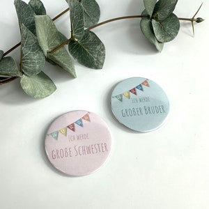 Könnte beinhalten: Zwei runde rosa und blaue Knöpfe mit bunten Flaggen und dem Text "Ich werde große Schwester" und "Ich werde großer Bruder".