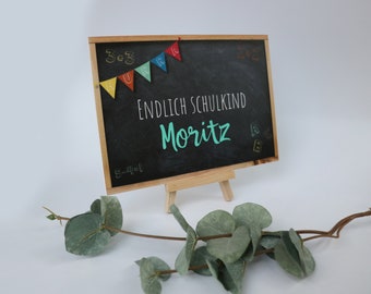 Puede incluir: Un pequeño caballete de madera con una pizarra negra en la que está escrito "ENDLICH SCHULKIND Moritz" con tiza blanca. Banderas de papel de colores con las letras "HURRA" están enhebradas en la parte superior de la pizarra. La pizarra está decorada con letras y números de colores.