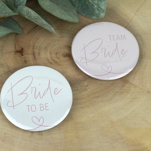 Könnte beinhalten: Zwei runde weiße und rosa Knöpfe mit dem Text "Bride to Be" und "Team Bride" in rosa Schrift. Beide Knöpfe haben ein kleines rosa Herz.