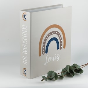 Könnte beinhalten: Ein beiger Ordner mit einem Regenbogen-Design in den Farben Braun, Orange, Blau und Grau. Der Regenbogen hat weiße Punkte. Der Name "Louis" ist in Weiß unter dem Regenbogen gedruckt. Der Text "Ihr Wunschlexi" ist auf dem Rücken des Ordners gedruckt.