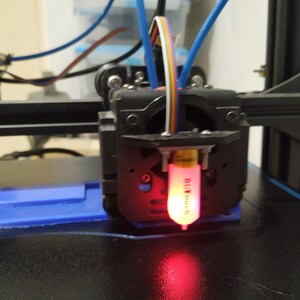 May include: Close-up of a 3D printer nozzle with a red filament inserted. The nozzle is attached to a black metal frame with a fan and wires.