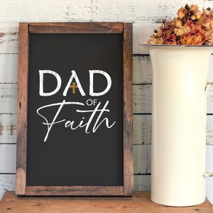 May include: A black chalkboard sign with a wooden frame. The sign says "DAD OF Faith" with a gold cross between the "D" and the "A".