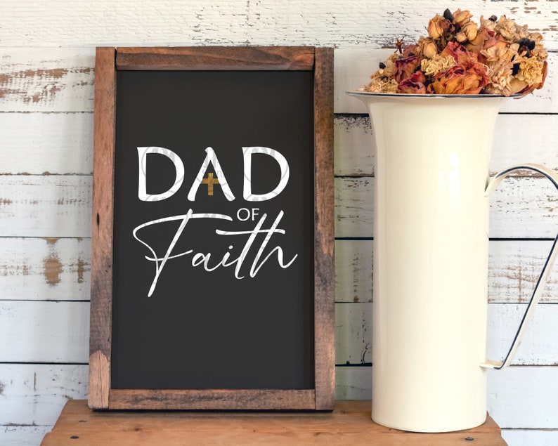May include: A black chalkboard sign with a wooden frame. The sign says "DAD OF Faith" with a gold cross between the "D" and the "A".