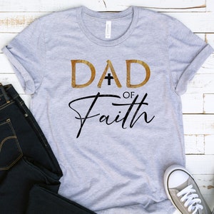 May include: A gray t-shirt with the text "DAD of Faith" in gold and black lettering. The word "DAD" has a cross in the middle of the "A".