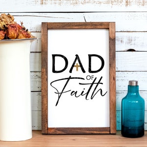 May include: A framed sign with a white background and black text that reads "DAD of Faith" with a gold cross in the "A".