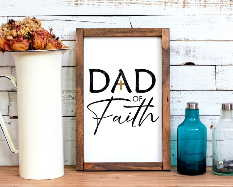 May include: A framed sign with a white background and black text that reads "DAD of Faith" with a gold cross in the "A".