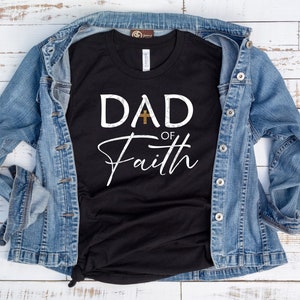 May include: Black t-shirt with white text that reads "DAD of Faith" with a gold cross between the words "DAD" and "of". The shirt is laid on a white wooden surface with a blue denim jacket draped over it.