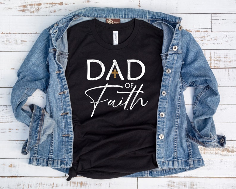 May include: Black t-shirt with white text that reads "DAD of Faith" with a gold cross between the words "DAD" and "of". The shirt is laid on a white wooden surface with a blue denim jacket draped over it.