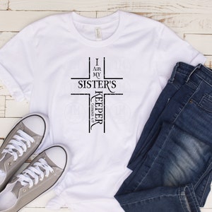 May include: White t-shirt with a black cross graphic that says "I am my sister's keeper, Genesis 4:9". The shirt is laid out on a white wooden surface with a pair of gray sneakers and blue jeans.