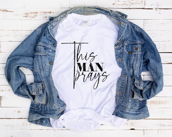 Puede incluir: Camiseta blanca con texto negro que dice "This Man Prays". La camiseta está doblada y colocada sobre una superficie de madera blanca con una chaqueta vaquera azul drapeada sobre ella.