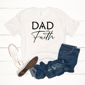 May include: A white t-shirt with the text "DAD OF Faith" in black lettering. The shirt is laid flat on a white wooden surface with a pair of white sneakers and blue jeans.