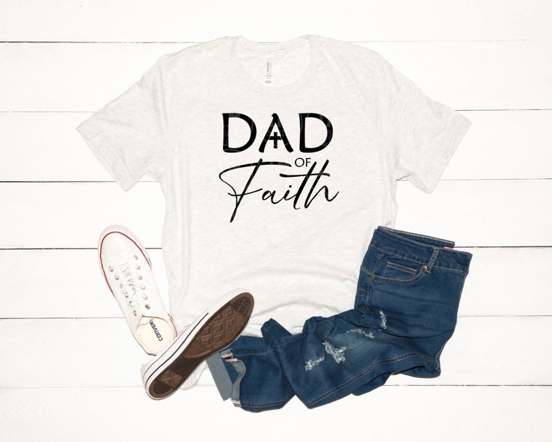 May include: A white t-shirt with the text "DAD OF Faith" in black lettering. The shirt is laid flat on a white wooden surface with a pair of white sneakers and blue jeans.