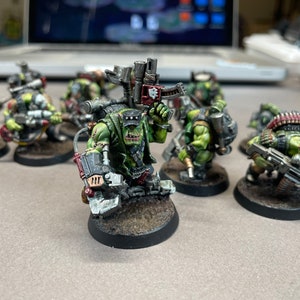 May include: A collection of miniature green-skinned Orc figures, each armed with various weapons and gear. The figures are painted with detailed features and stand on small, oval bases. The scene suggests a tabletop war game or hobby project.