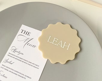 May include: A beige place card with the name "LEAH" printed in white. The place card is on a gray plate with a white menu card that says "The Menu" and lists appetizers, salad, and entrees.
