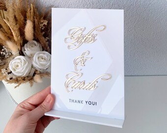Op de afbeelding: Een wit bord met gouden letters met de tekst "Gifts & Cards" en de woorden "Thank You!" onderaan.