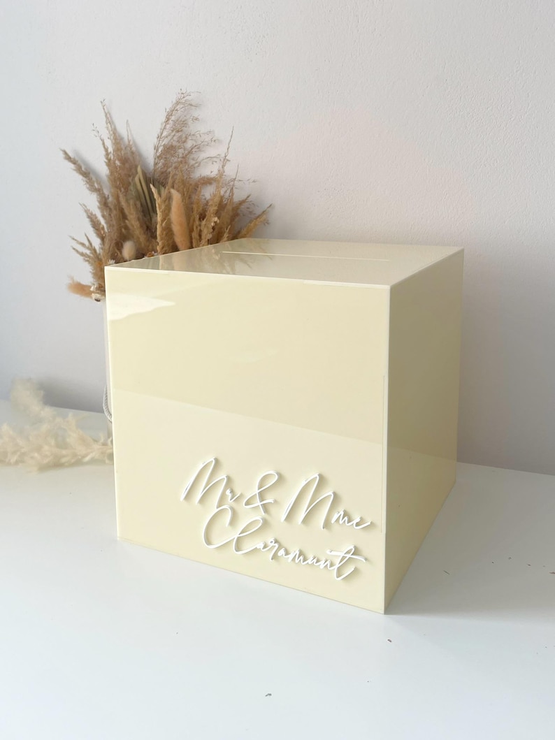 Op de afbeelding: Een lichtgele, vierkante trouwkaartendoos met de woorden "Mr. & Mrs. Clermont" in witte letters. De doos is glad en glanzend. Gedroogde grassen zijn zichtbaar op de achtergrond.