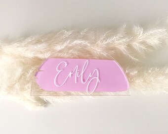 May include: A clear acrylic place card with a pink painted half-circle and the name "Emily" written in white script.
