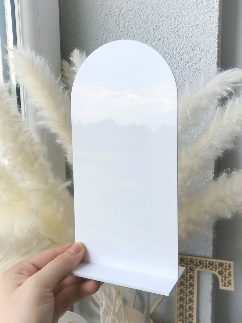 May include: White acrylic display stand with an arched top and a flat base. The stand is designed to hold small items, such as cards, photos, or jewelry.