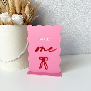 Könnte beinhalten: Rosa Tischnummernschild mit dem Wort "TABLE" und der Zahl "eins" in einer kursiven Schrift. Unter der Zahl befindet sich eine rote Schleife.