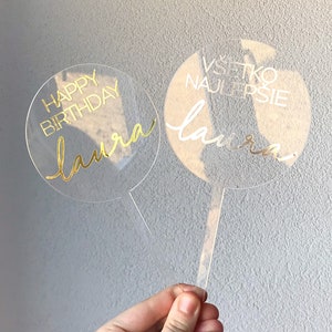 May include: Two clear acrylic cake toppers with gold lettering. The first topper says "Happy Birthday Laura" and the second topper says "Vsetko Najlepsie Laura".