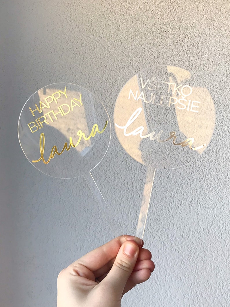 May include: Two clear acrylic cake toppers with gold lettering. The first topper says "Happy Birthday Laura" and the second topper says "Vsetko Najlepsie Laura".