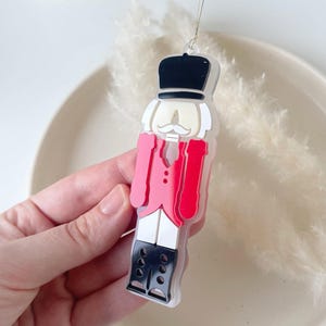 Könnte beinhalten: Ein Weihnachtsornament in Form eines Spielzeugsoldaten. Es hat einen schwarzen Hut, einen weißen Schnurrbart und eine rote Jacke. Das Ornament hat schwarze Stiefel und eine weiße Hose, die an einem silbernen Faden aufgehängt sind.