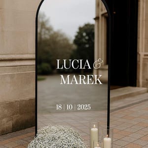 以下が含まれることがあります： 黒いフレームのアーチ型ミラーには「Lucia & Marek」の名前と日付「18 | 10 | 2025」が表示されています。 ガラスシリンダーに入ったキャンドルと白い花のクラスターがあり、結婚式やイベントの装飾のようです。