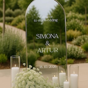 May include: A reflective wedding sign with the words "Welcome to our wedding", the names "SIMONA & ARTUR", and the date "18. 10. 2026". White candles in glass holders and white flowers are arranged in front of the sign.