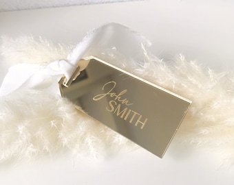 May include: A gold mirror tag with the name "John Smith" engraved in a cursive font. The tag is tied with a white ribbon and placed on a white fluffy surface.