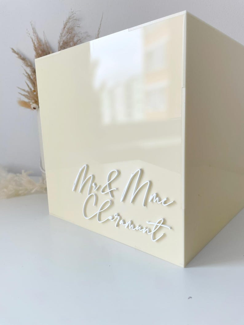Op de afbeelding: Een cr&egrave;mekleurige vierkante doos met de woorden "Mr. & Mrs. Claremont" in witte letters. De doos heeft een glanzende afwerking, waarschijnlijk voor een bruiloft of speciale gelegenheid.