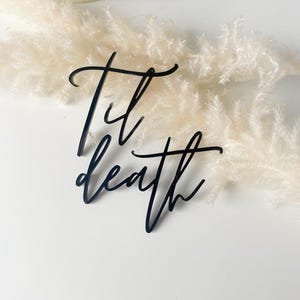 Puede incluir: Un letrero de acrílico negro con las palabras "Til death" escritas en cursiva.