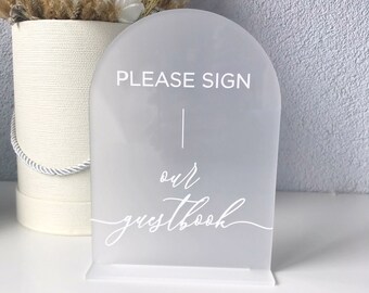 Op de afbeelding: Wit acryl bord met afgeronde bovenkant, staand op een witte voet. Het bord heeft de tekst "PLEASE SIGN our guestbook" in witte letters.