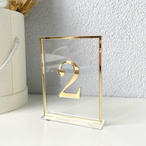 Puede incluir: Un porta números de mesa de acrílico transparente con marco dorado y el número "2" en letras doradas.