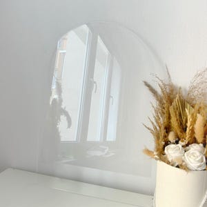 Könnte beinhalten: Eine transparente, bogenförmige Acrylplatte steht auf einer weißen Oberfläche und spiegelt ein Fenster wider. Ein runder, weißer Behälter enthält einen Strauß aus getrockneten Gräsern und weißen Rosen, der der Szene eine natürliche Note verleiht.