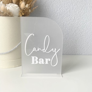 Puede incluir: Un letrero de acrílico blanco con bordes redondeados que dice "Candy Bar" en letras blancas en cursiva con la palabra "Bar" en letras blancas en negrita.