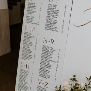 Könnte beinhalten: Ein Hochzeits-Sitzplan in Weiß und Gold, mit alphabetisch angeordneten Namen. Die goldene Schrift befindet sich auf weißem Hintergrund. Ein kleiner Strauß weißer Blumen befindet sich unten.