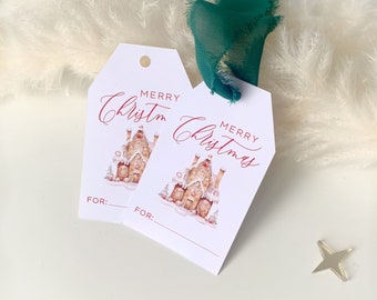 Könnte beinhalten: Zwei weiße Geschenkanhänger mit einem Aquarell-Lebkuchenhaus-Design und den Worten "MERRY Christmas" in roter Schrift. Ein dunkelgrünes Band ist an den Anhängern befestigt. Die Anhänger haben einen Platz für "FOR:" und ein kleiner goldener Stern im Hintergrund.