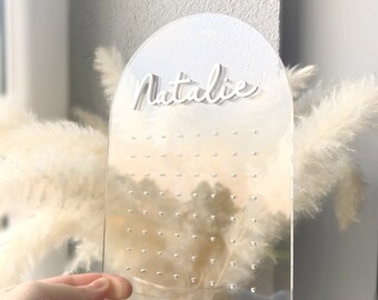 May include: Clear acrylic earring display with an arched top and a base. The name "Natalie" is in white script across the top. The display has multiple small holes for earrings. A hand is holding the display.