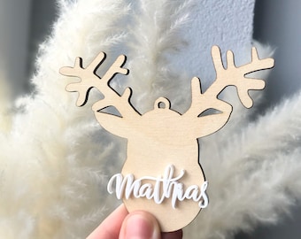 May include: A wooden reindeer head ornament with the name "Mathias" in white script. The reindeer head is light brown with detailed antlers. The ornament has a small hole for hanging. The background features fluffy white pampas grass.